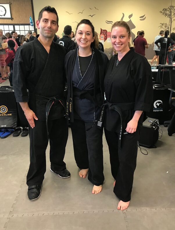 Three martial artists posing indoors, two wearing black gi and one in casual clothes.